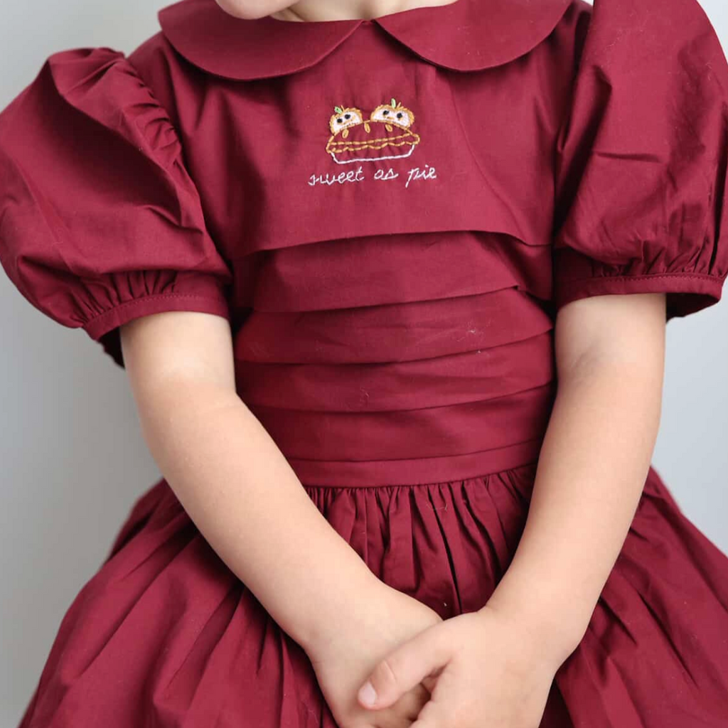 Child wearing a burgundy dress with a embroidered design on a plain background